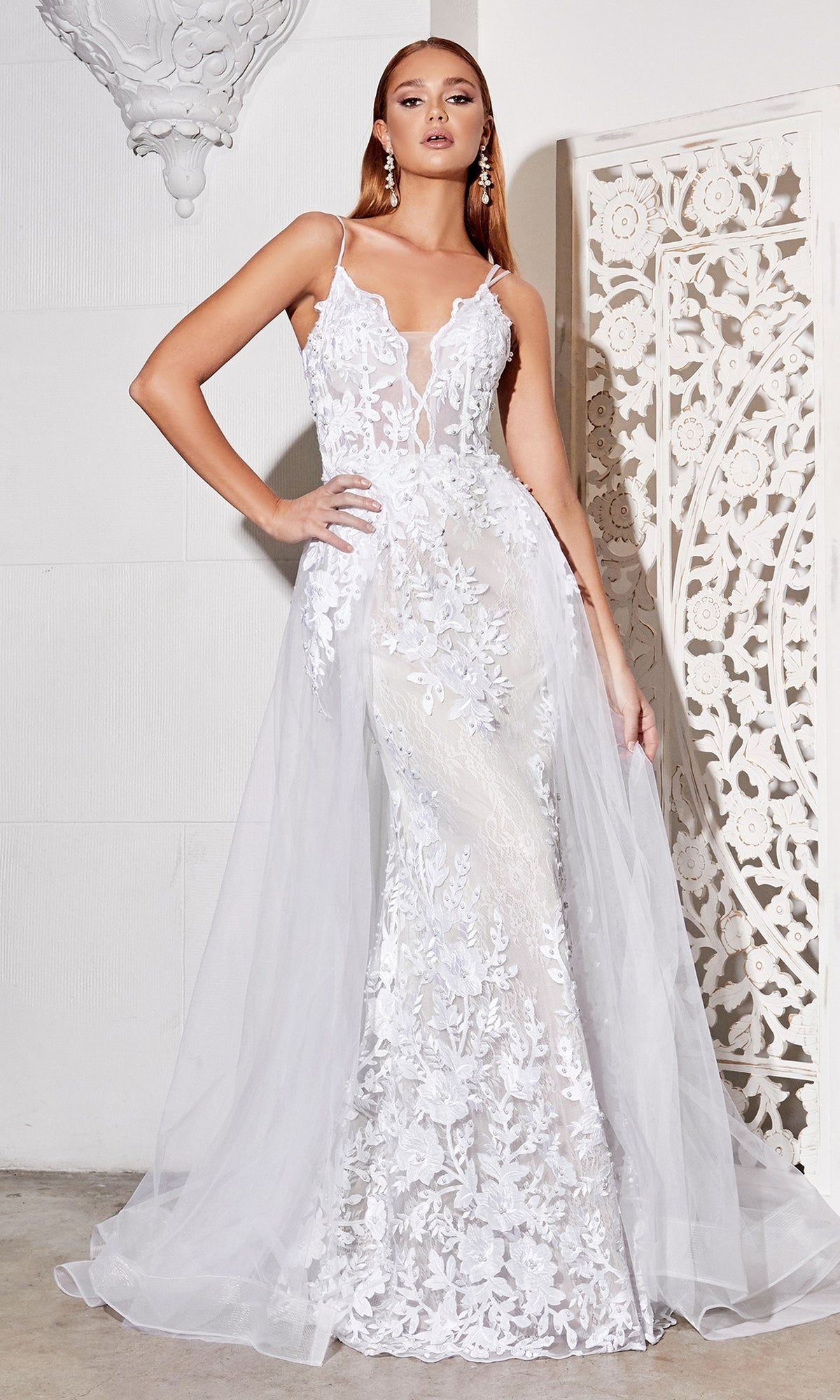 A woman stands indoors against a white decorative backdrop wearing the Sheer-Corset Long White Lace Wedding Dress CD931W, a fitted sleeveless bridal gown with an off-white sheer overlay and delicate floral appliqué details.